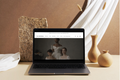 Laptop on a table with a website displayed, surrounded by decorative vases and a textured pillow.